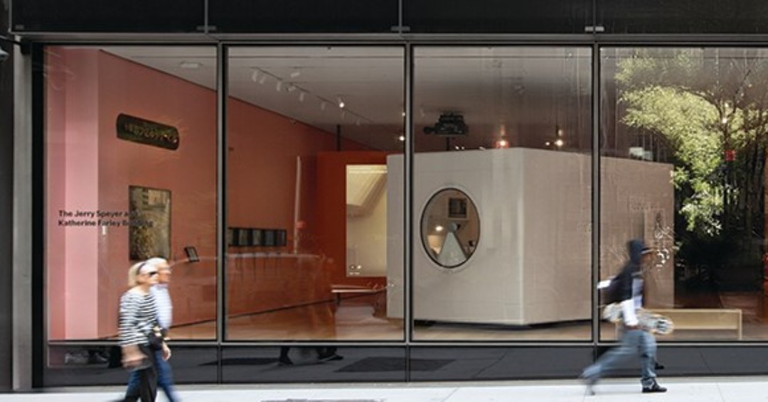 MoMA’s restored Nakagin capsule sits behind a street window beside pink exhibition graphics.