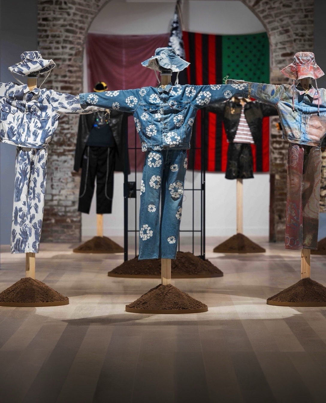 Three Denim Tears outfits on wooden crossbar stands above soil, with cotton wreath denim in the centre of a brick gallery.