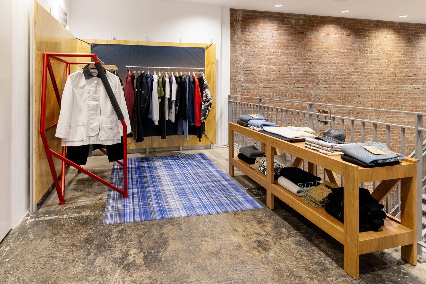 Stüssy clothing around a wooden table and red metal rail in a plywood space with a blue checked rug at DSMNY.