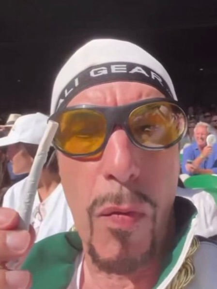 Sacha Baron Cohen in character as Ali G at the Wimbledon 2026 men's singles final, wearing a green and white tracksuit, gold chain and yellow tinted sunglasses.