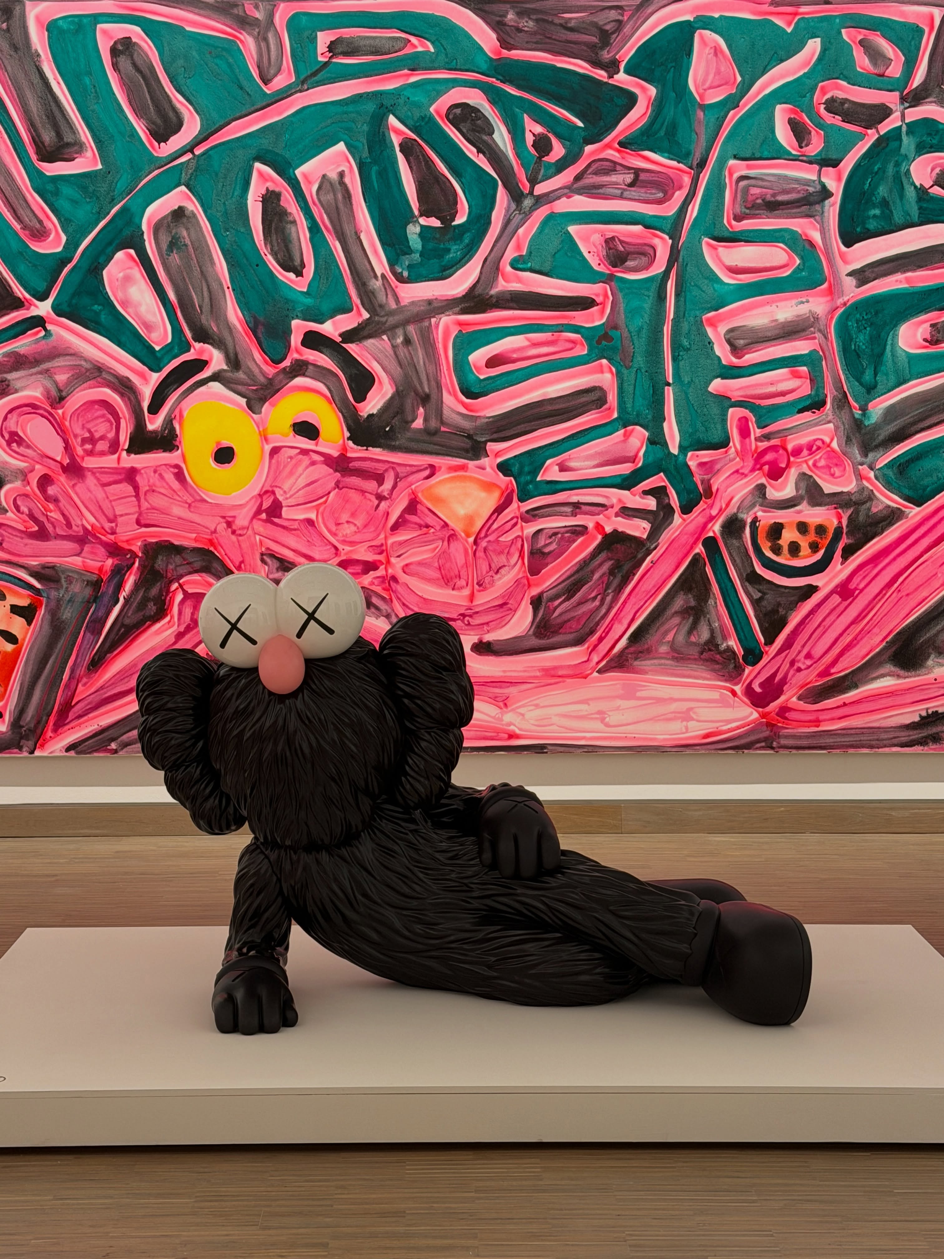 KAWS TIME OFF reclines in a gallery with a Pink Panther painting behind it.