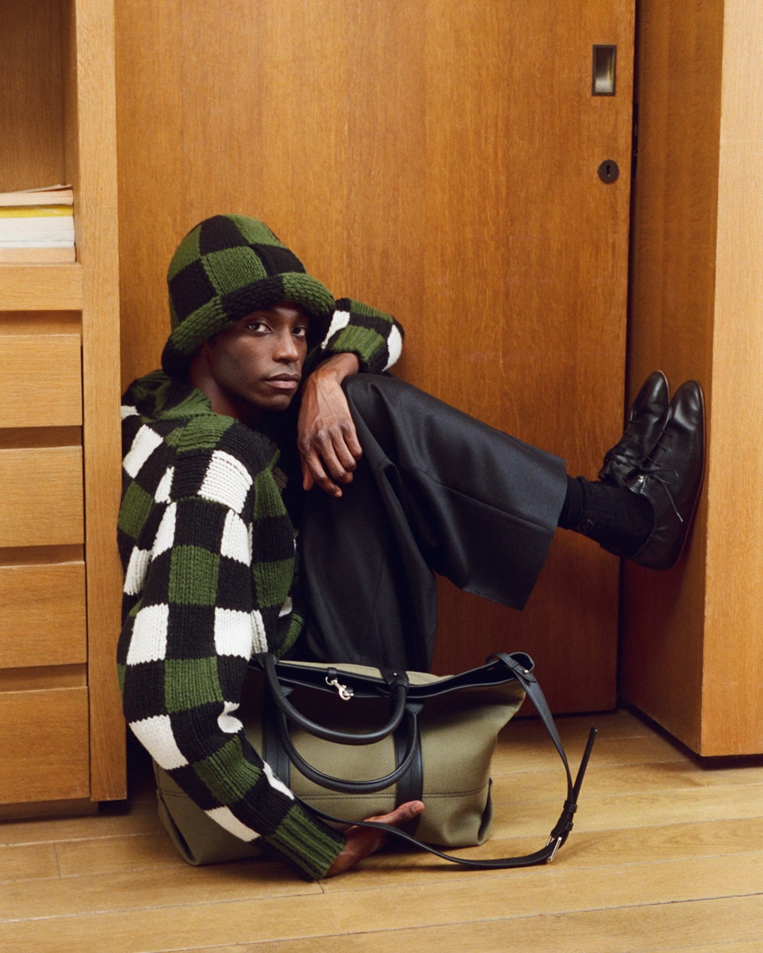 A Kenzo model wears an olive and dark checkerboard knit beside a wooden doorway.