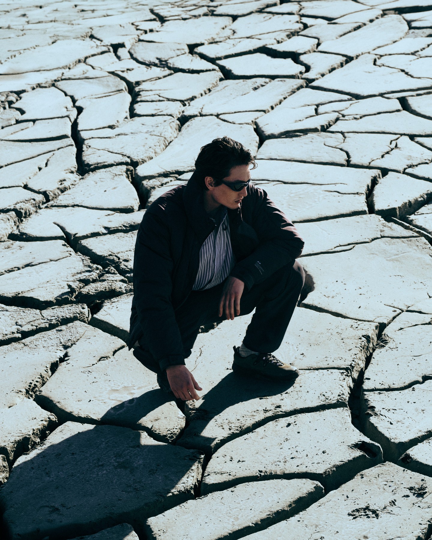A model crouches on pale cracked ground in dark Oakley outerwear and trousers with wraparound sunglasses.