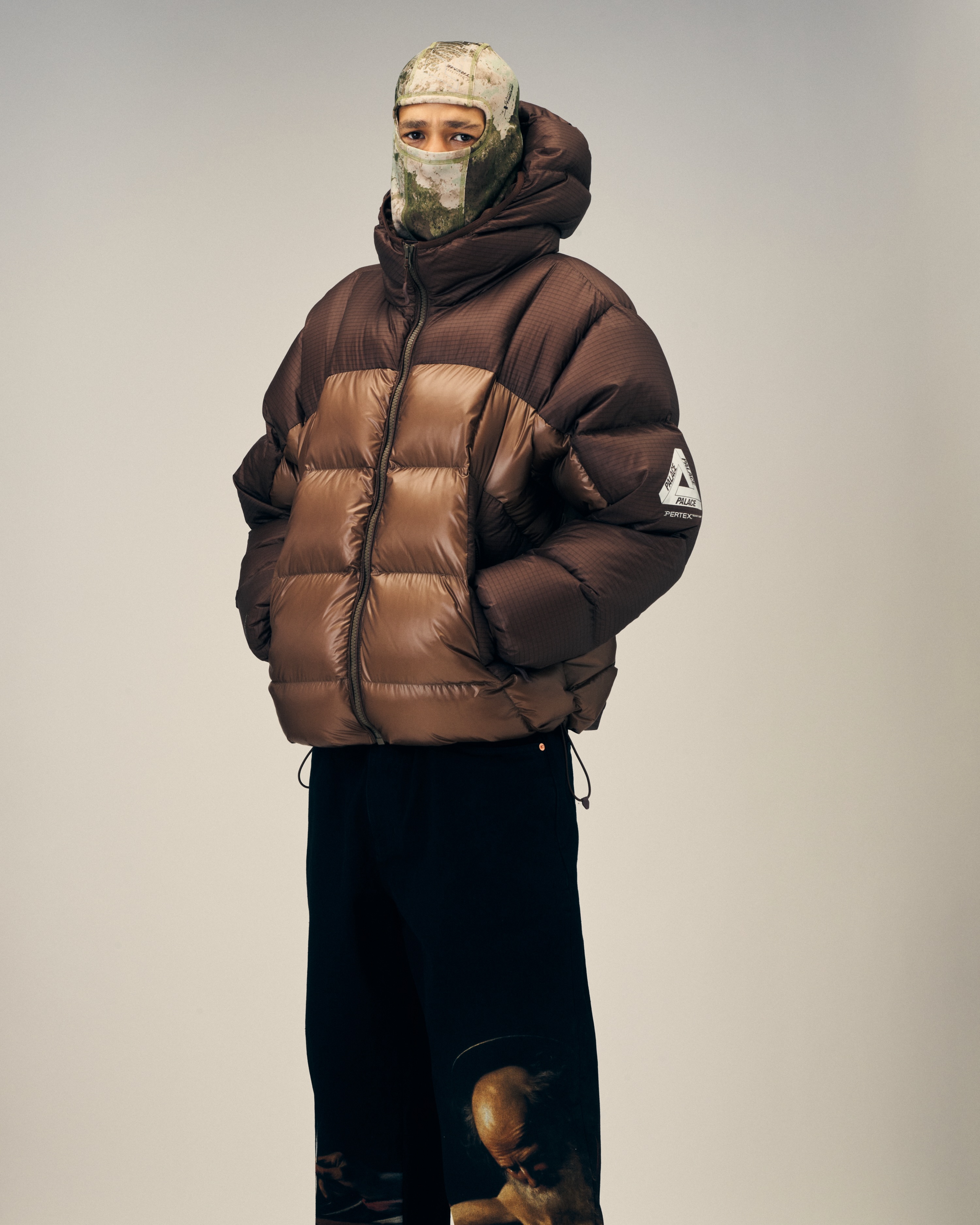 A model in a tan and dark brown hooded Palace puffer with Pertex sleeve branding, camouflage face covering and wide black trousers.