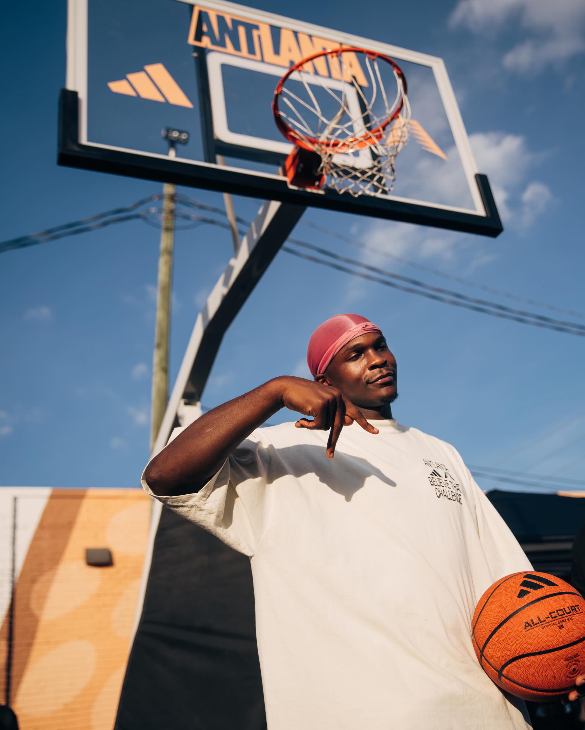 Anthony Edwards in a pink durag and cream Believe That Challenge shirt holding a basketball beneath an Antlanta backboard.