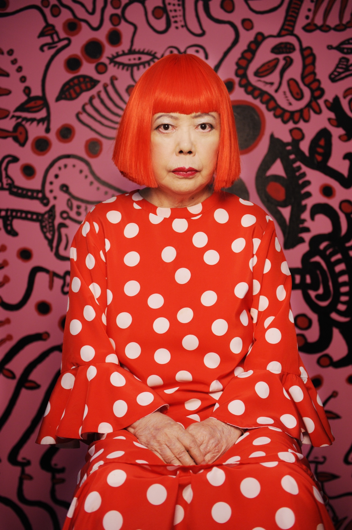 Yayoi Kusama photographed in a red and white polka dot dress and red bob wig against a patterned background