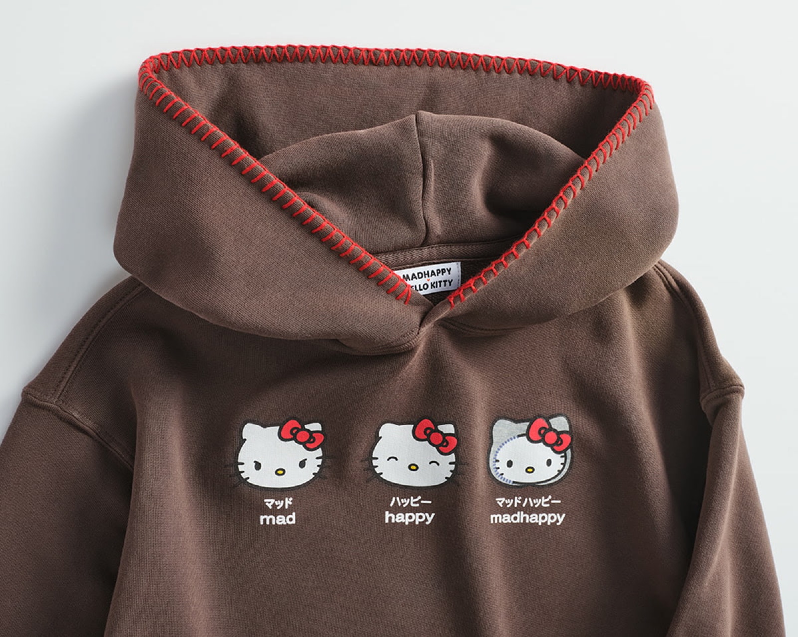 Brown Madhappy x Hello Kitty Emotions hoodie with three Hello Kitty faces reading mad, happy, and madhappy, and a red contrast stitch on the hood