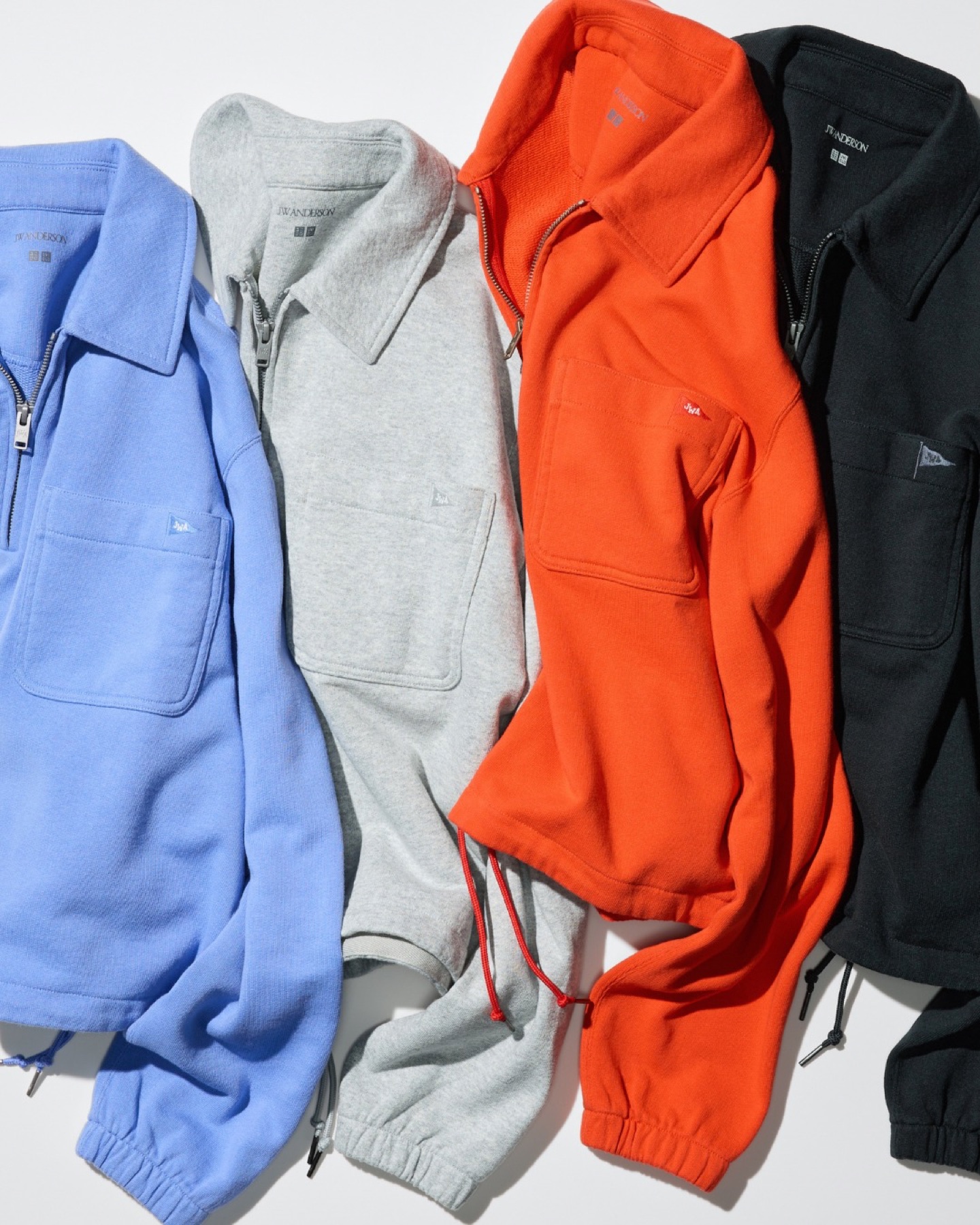 Four Uniqlo and JW Anderson half zip sweat pullovers in sky blue, grey, orange and black laid flat, each with a small JWA pennant on the chest pocket