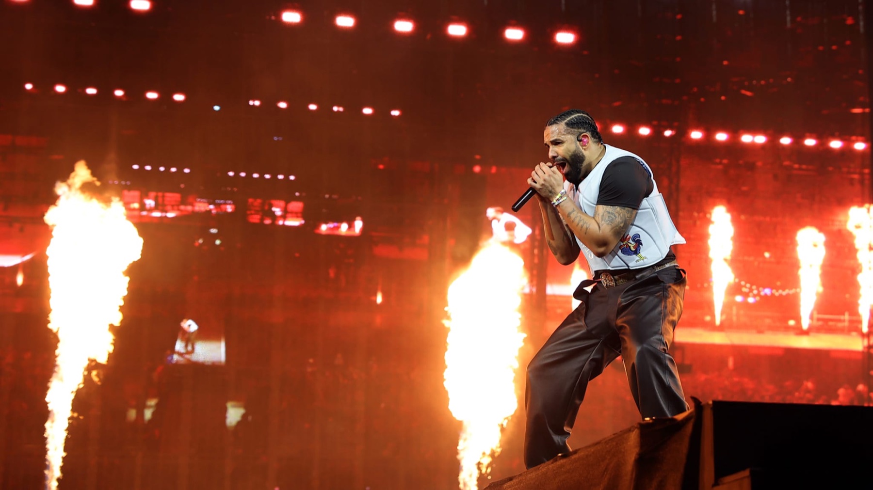 Drake performing live on stage under red pyrotechnic flames, microphone in hand