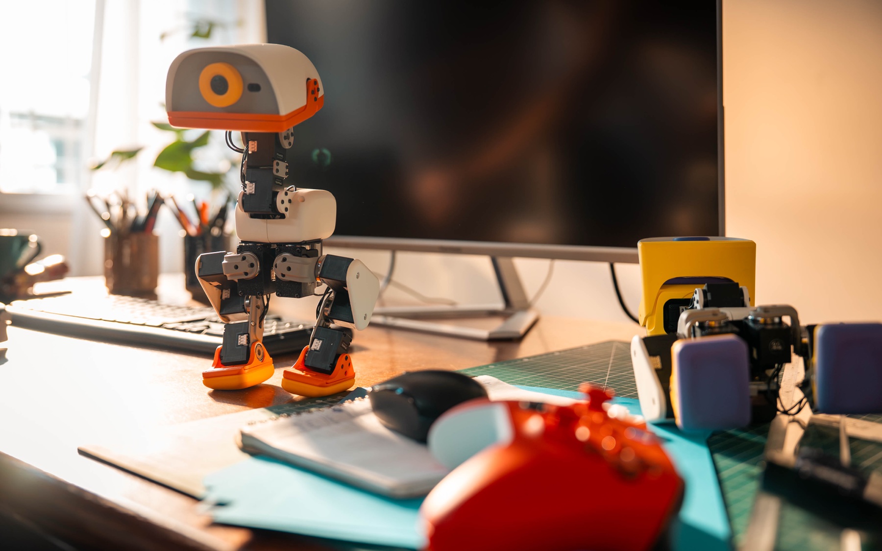 The Hugging Face Microduck, a small white and orange bipedal robot with a duck bill head and camera eye, standing on a desk